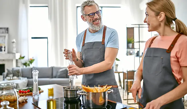 smiling middle age couple cooking in kitchen with aprons